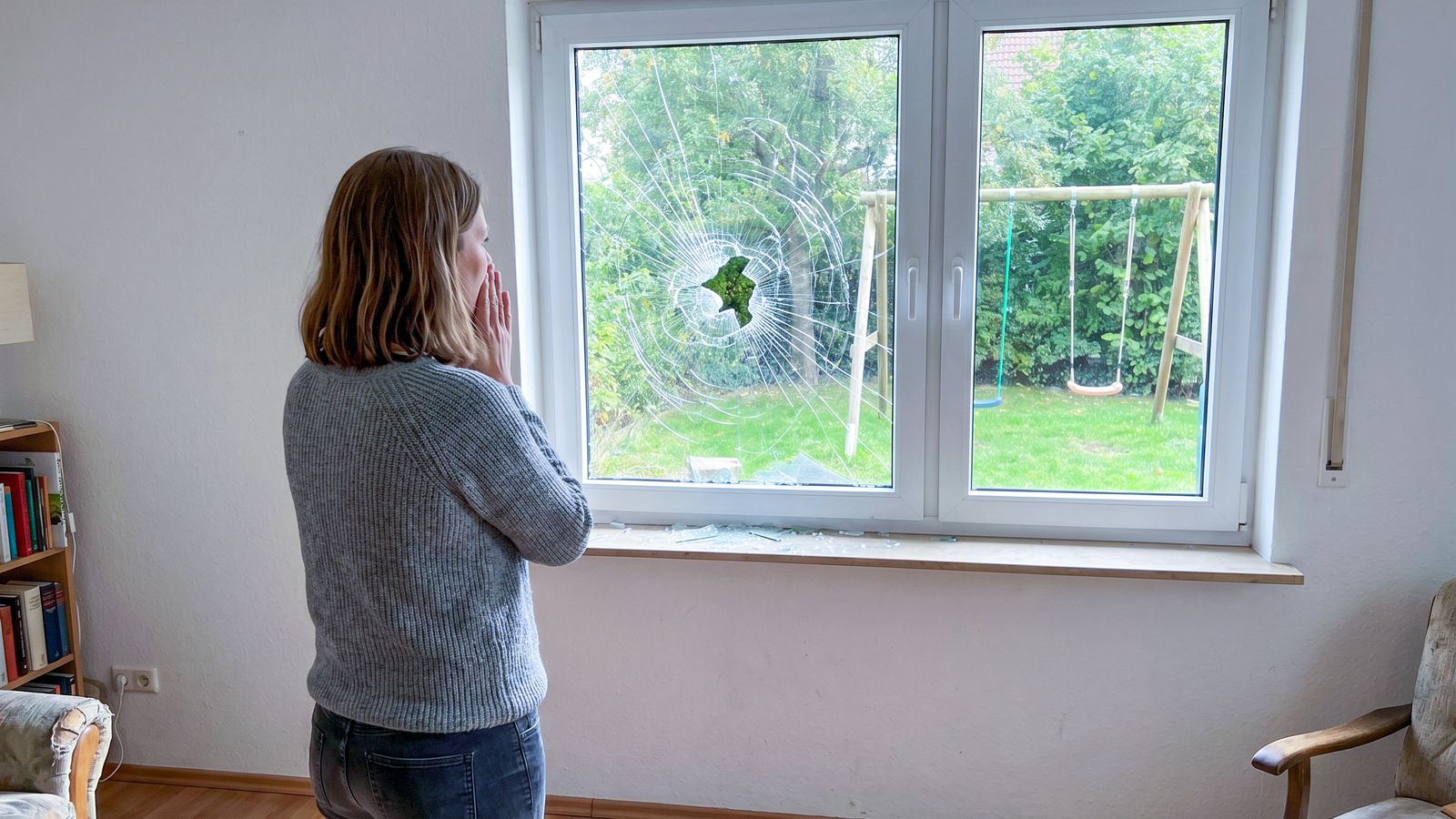 Erschrockene Frau steht vor einem zerbrochenen Fenster mit Loch im Glas und Glassplittern auf dem Wohnzimmerboden